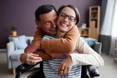 Partner umarmt seine Freundin, die im Rollstuhl sitzt, liebevoll von hinten