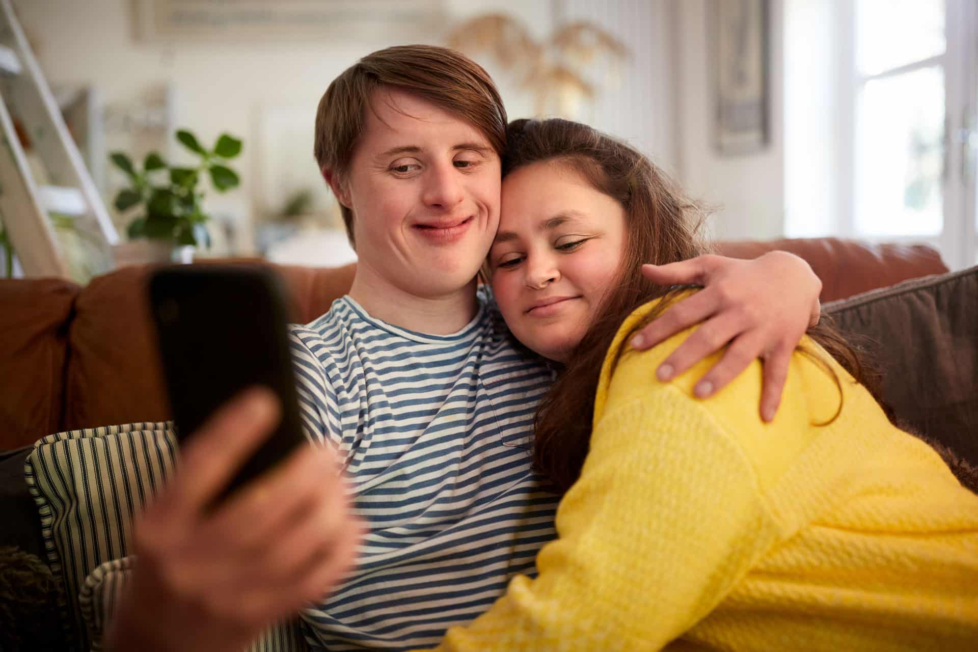 junges Pärchen mit Down-Syndrom auf Couch sitzend macht Selfie mit Handy