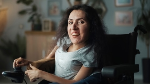 Lebensfrohe Frau mit spinaler Muskelatrophie in Rollstuhl sitzend, lächelt fröhlich direkt in Kamera